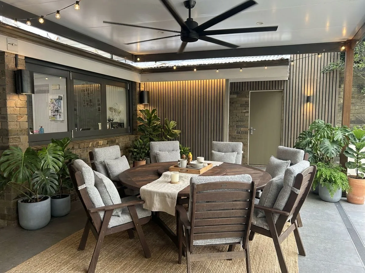 Transitional outdoor dining space with wood slat walls, dark wood table, light grey cushions, jute rug, string lights, photorealistic interior design photography, 8k